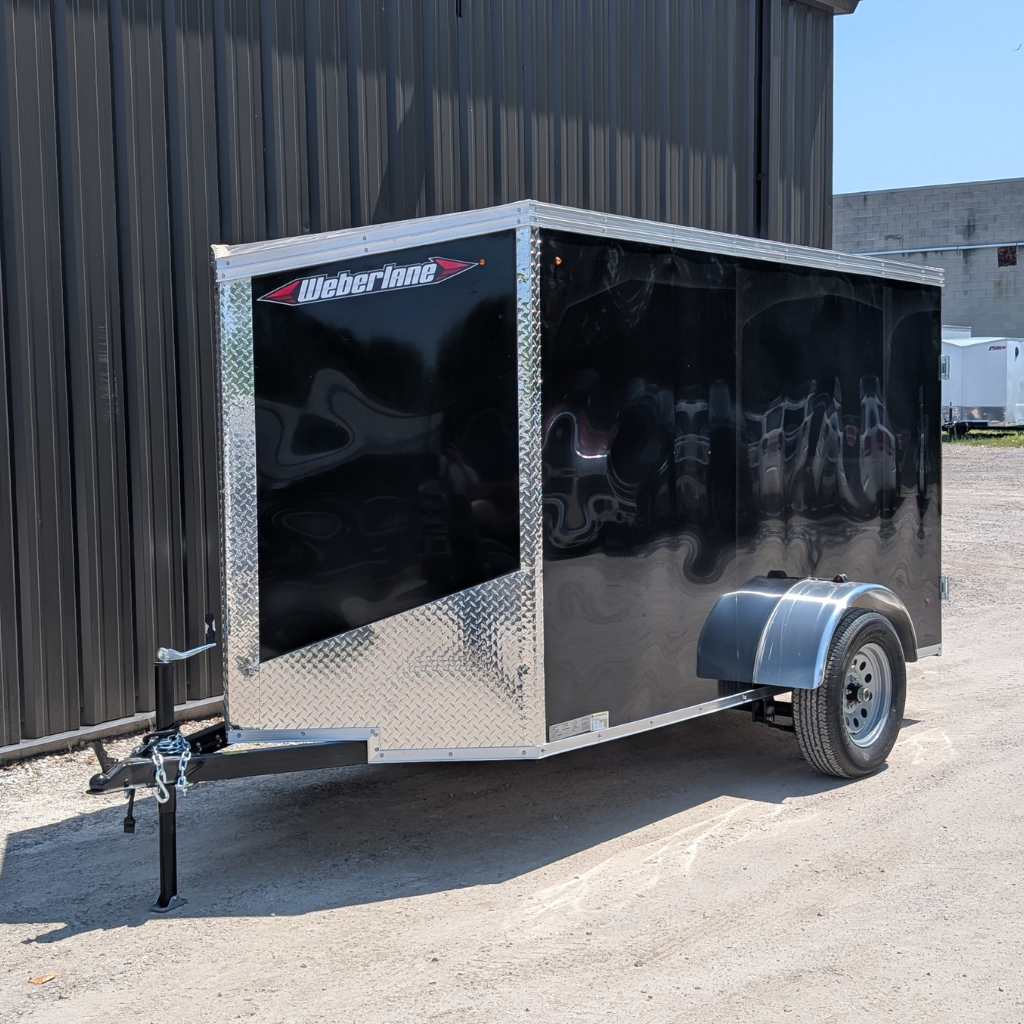 A 2025 WEBERLANE 5X10 ESTATE SERIES RAMP DOOR BLACK enclosed utility trailer is parked outside near a metal building on a sunny day.