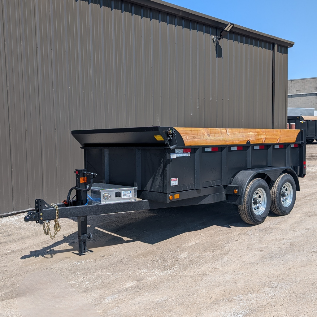 A 2026 CANADA TRAILER 6X12 DUMP 10,000 GVWR with black finish and wood sides is parked on a gravel lot beside a brown metal building.