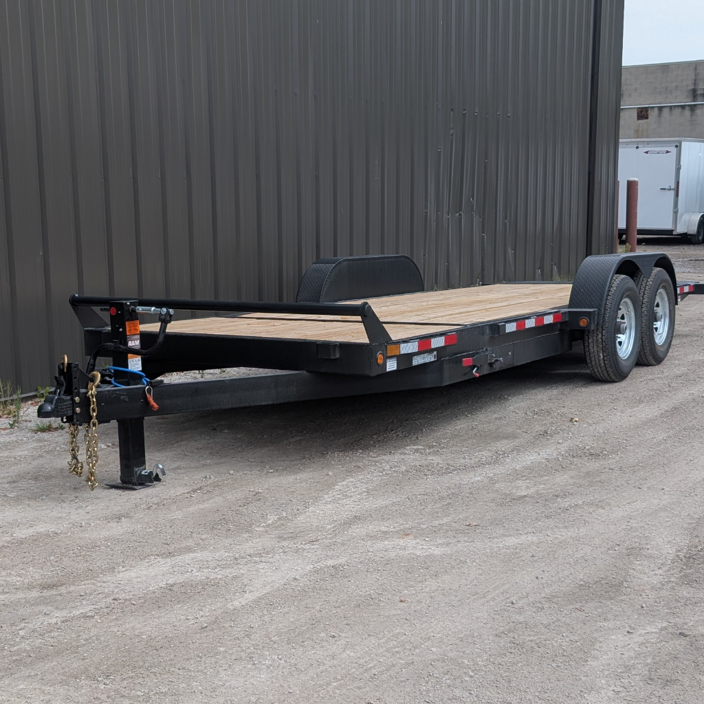 2026 CANADA TRAILER 7X21 Gravity Tilt 14,000 GVWR utility trailer with dual axles and wooden deck parked on gravel beside a corrugated metal building.