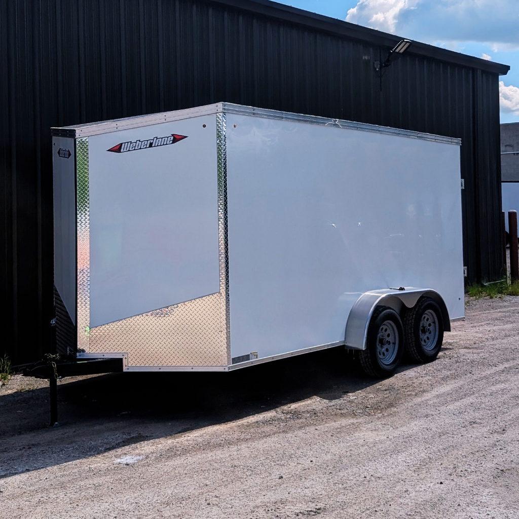 A 2026 WEBERLANE 7X16 Estate Series trailer with 12" extra height and a ramp door in white is parked on gravel beside a black metal building under partly cloudy skies.
