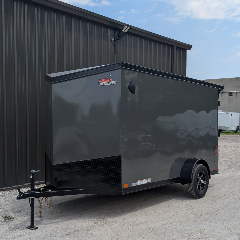 A 2025 UNITED TRAILER 6X12 FLAT TOP V-NOSE in battleship gray with a ramp door and 6.6’ height is parked on gravel beside a metal building under a partly cloudy sky.
