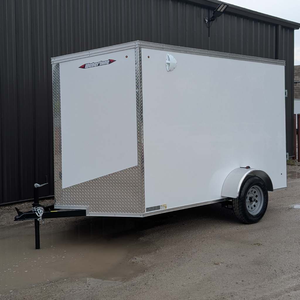 A 2026 CANADA TRAILER 6X12 steel side utility trailer with a 2990 GVWR and single axle is parked on a muddy gravel surface beside a dark metal building.