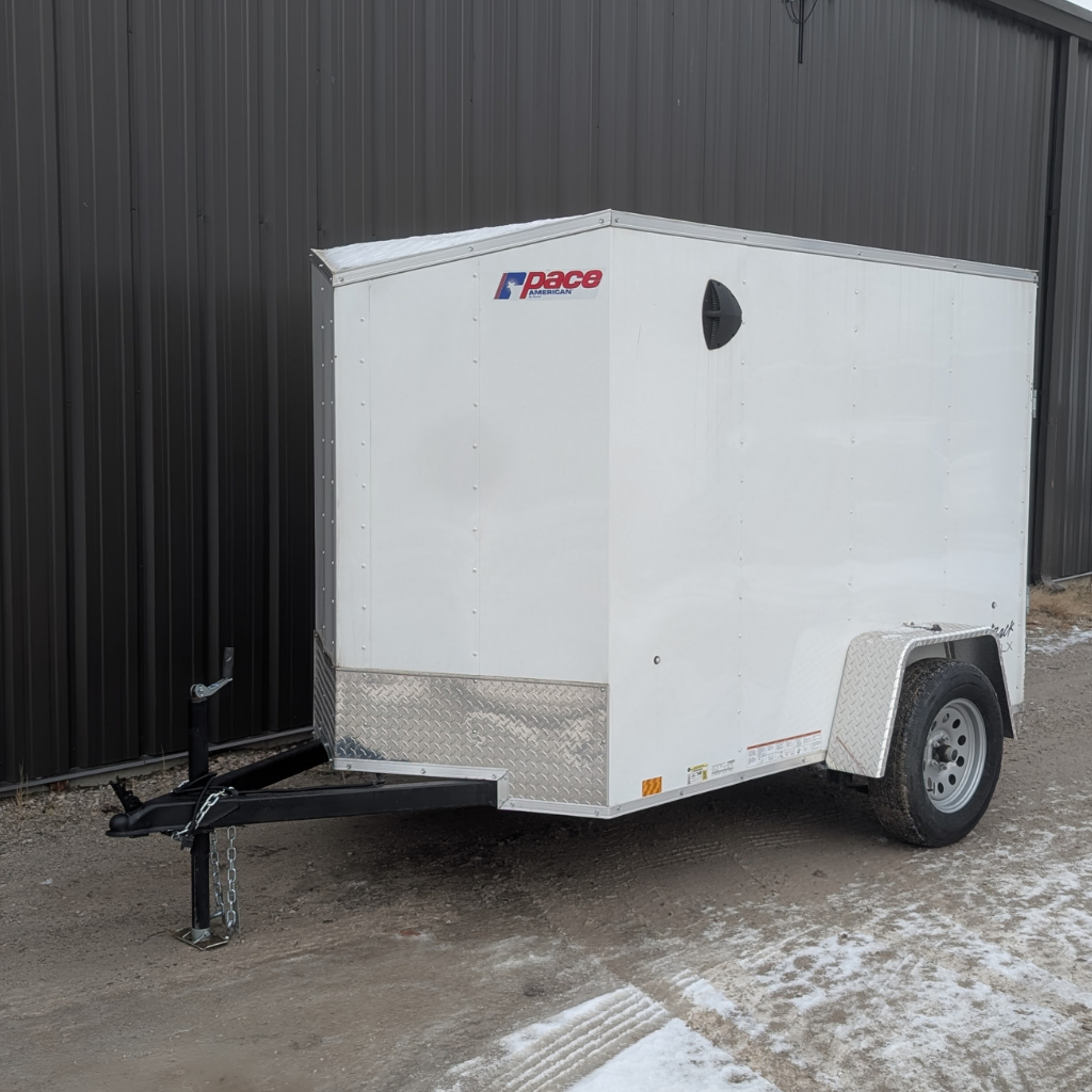 A 2023 PACE OB5X8SI2DLX Outback Flat Top enclosed cargo trailer with barn door and single axle in white is parked on gravel next to a corrugated metal building.