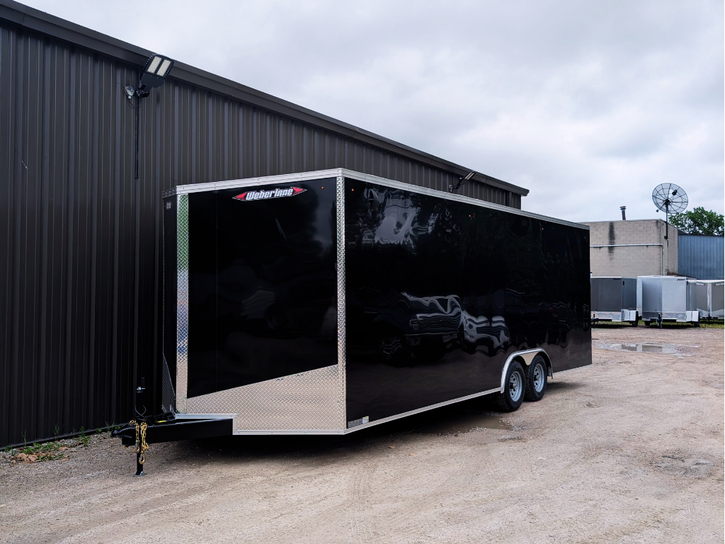A 2026 WEBERLANE 85X24 CONTRACTOR SERIES utility trailer with a 12" extra height ramp door, finished in black, is parked on a gravel lot beside a metal building under an overcast sky.