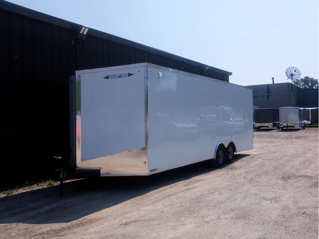 A 2026 WEBERLANE 85X24 CONTRACTOR SERIES trailer with 12" extra height, ramp door, and white exterior is parked on a gravel lot beside a black building under a sunny sky.