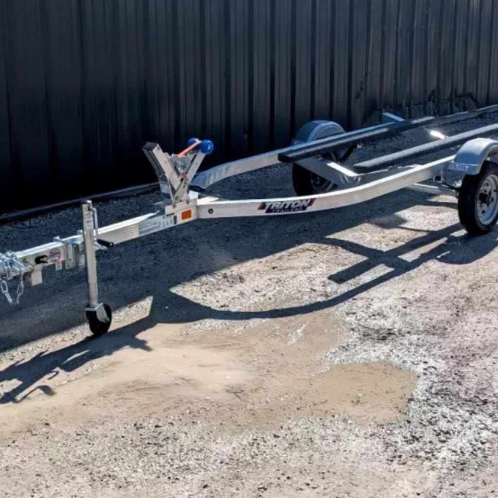 A 2025 TRITON SINGLE BED JETSKI TRAILER with a winch and wheel stand is parked on gravel beside a black metal building.