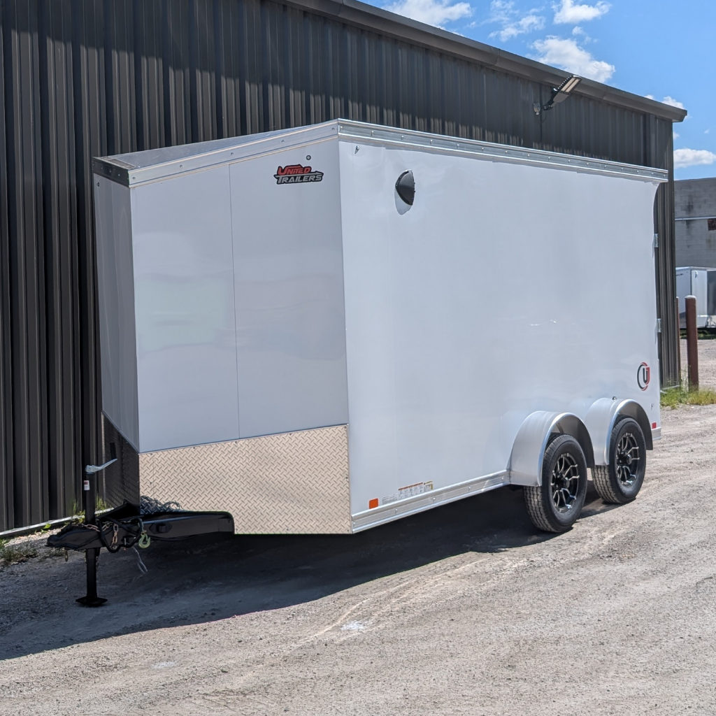A 2026 UNITED TRAILER 7X14 FLAT TOP V-NOSE with 12" extra height, ramp door, and white exterior is parked on gravel near a metal building under a partly cloudy sky.