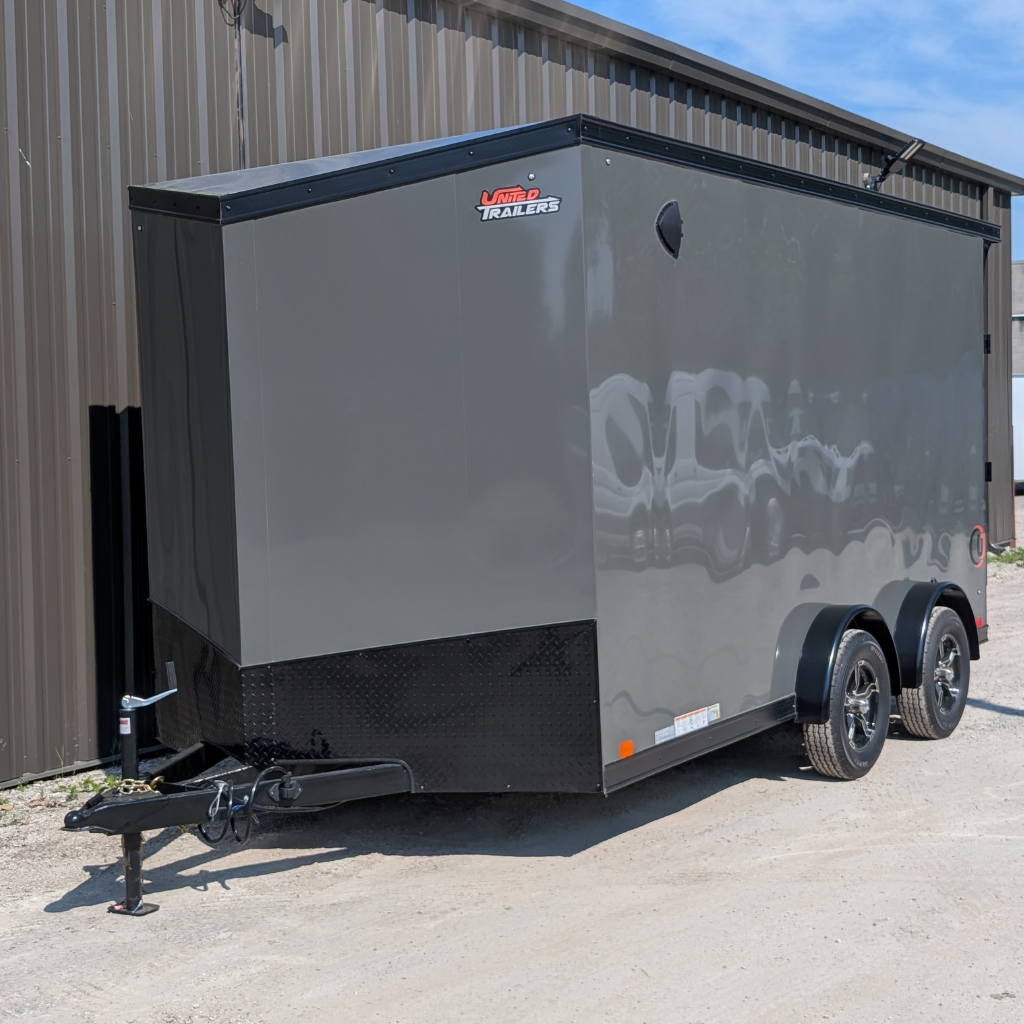 A 2025 UNITED TRAILER 75X14 FLAT TOP V-NOSE in battleship gray with dual axles and black wheels is parked on gravel beside a metal building.
