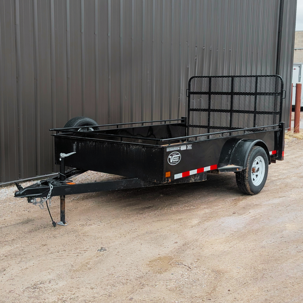 A USED 2021 CANADA TRAILER 6X10 Steel Side Utility Trailer (2990 GVWR) with a mesh ramp gate and single axle is parked on gravel next to a metal building.