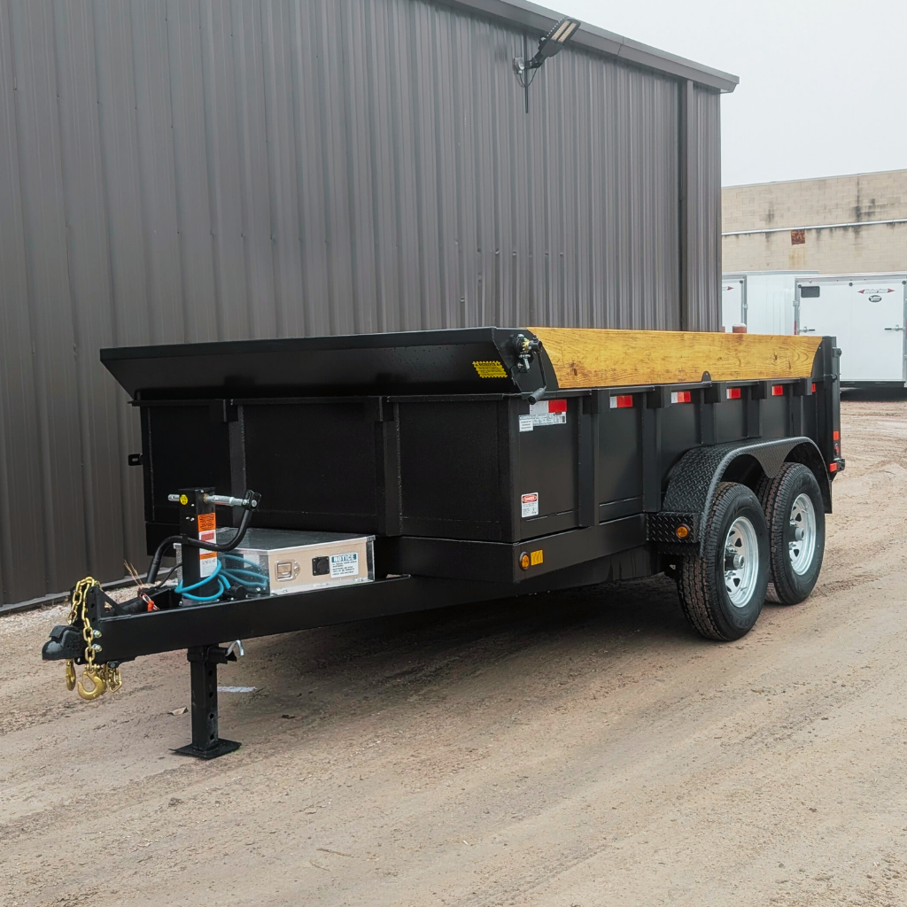 A 2026 CANADA TRAILER 83X12 DUMP 14000 GVWR with wooden side rails is parked on a gravel surface near a metal building.
