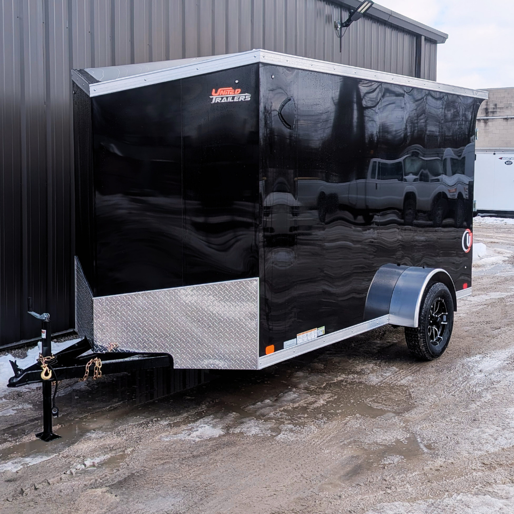 2026 UNITED TRAILER 6X12 FLAT TOP V-NOSE, 6.6' height, black with ramp door and diamond plate trim, single axle, parked on wet slush beside a dark metal building.