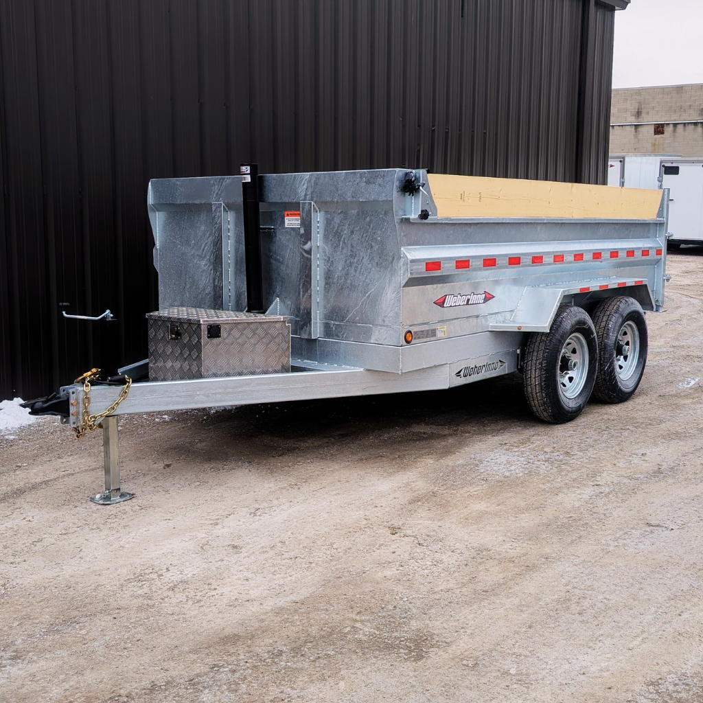 A 2026 WEBER LANE 8X12 CONTRACTOR SERIES 14000 GVWR galvanized dump trailer is parked on gravel beside a black metal building.