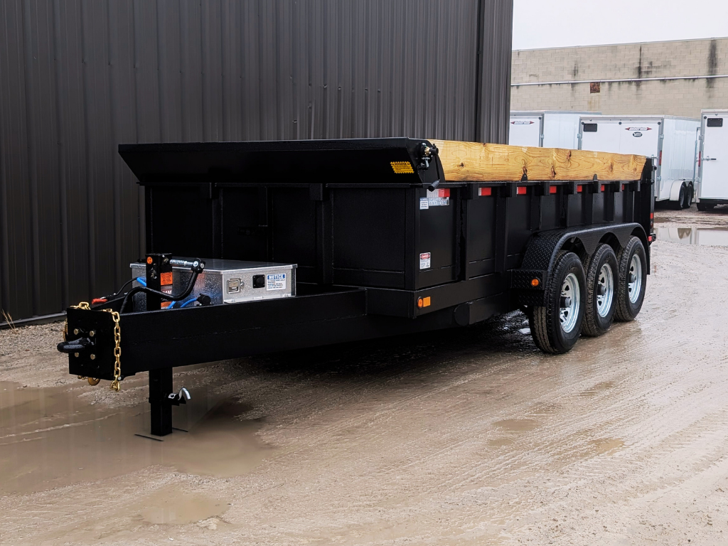 A 2026 Canada Trailer 83x16 Ultra Series Dump (21,000 GVWR), black with triple axles and wooden side extensions, is parked on wet gravel next to a metal building.