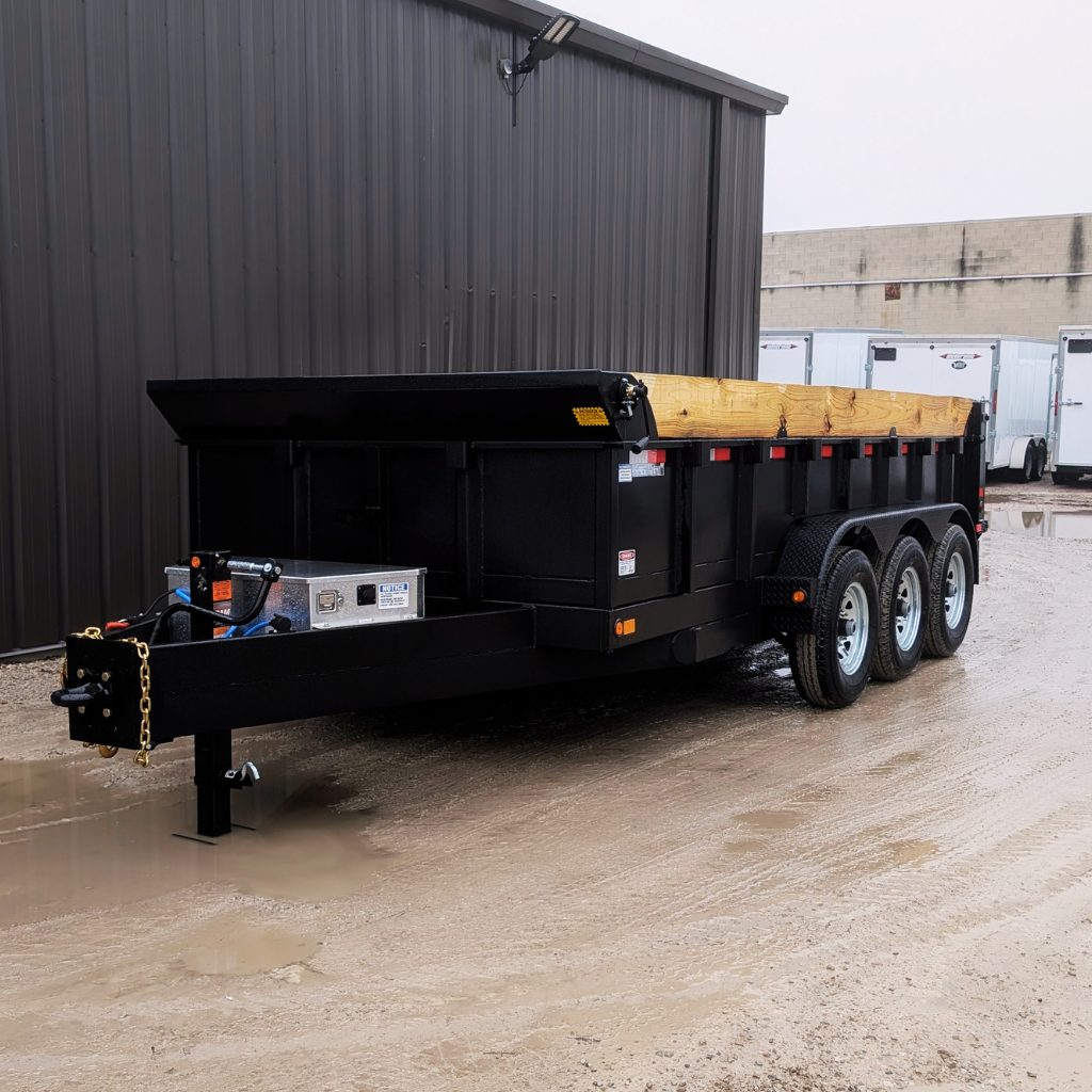A 2026 Canada Trailer 83X16 Ultra Series Dump 21,000 GVRW with black triple axles and wooden side extensions is parked on wet ground near a metal building and several white trailers.
