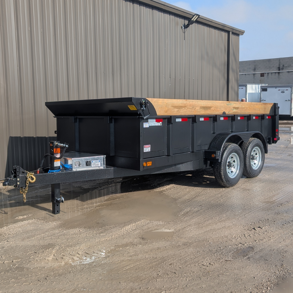 A 2026 CANADA TRAILER 83X14 DUMP 14,000 GVWR with black tandem axles and wooden side extensions is parked on muddy gravel beside a metal building.