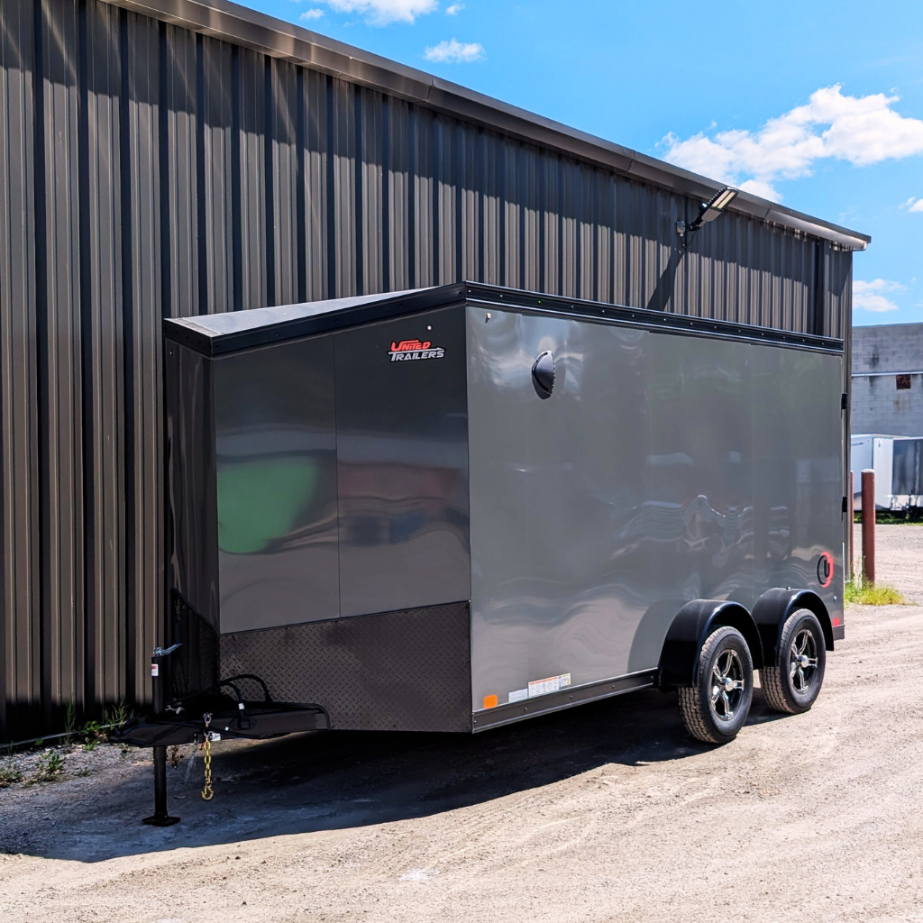 A 2026 United Trailer 7x16 Flat Top V-Nose Battleship Gray enclosed cargo trailer with a ramp door is parked on gravel beside a metal-sided building on a sunny day.