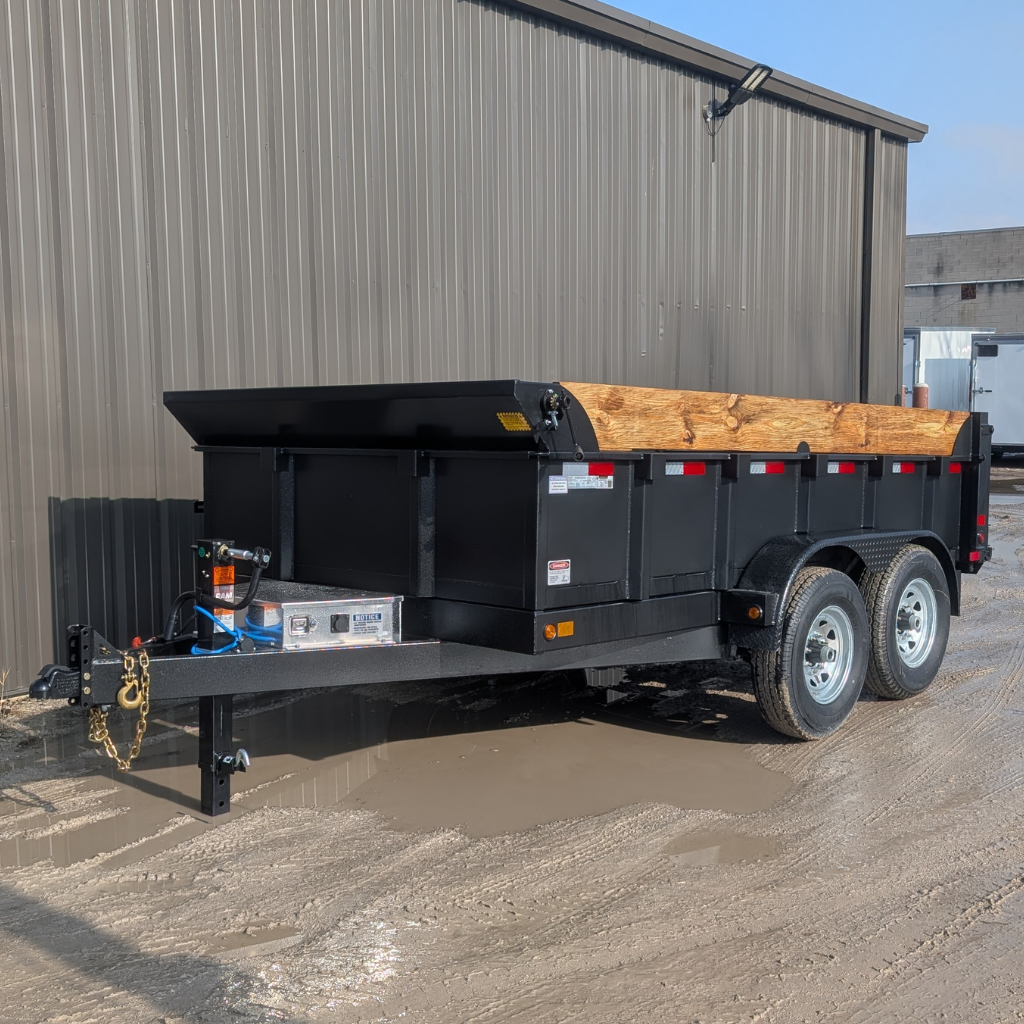 A 2026 CANADA TRAILER 83X12 DUMP 14000 GVWR with wooden side extensions is parked on a muddy surface beside a metal building.