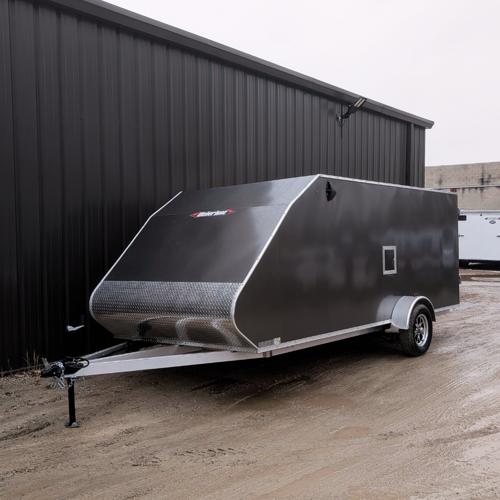 A grey 2026 Weberlane 7x16 Aluminum Snow Series enclosed utility trailer with a ramp door and single axle is parked on a muddy surface next to a metal-sided building.