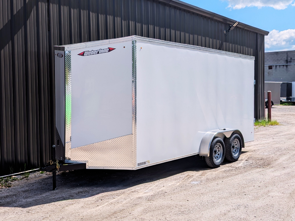 A 2026 WEBERLANE 7X16 Estate Series white enclosed trailer with 6" extra height and ramp door is parked on gravel beside a dark metal-sided building.