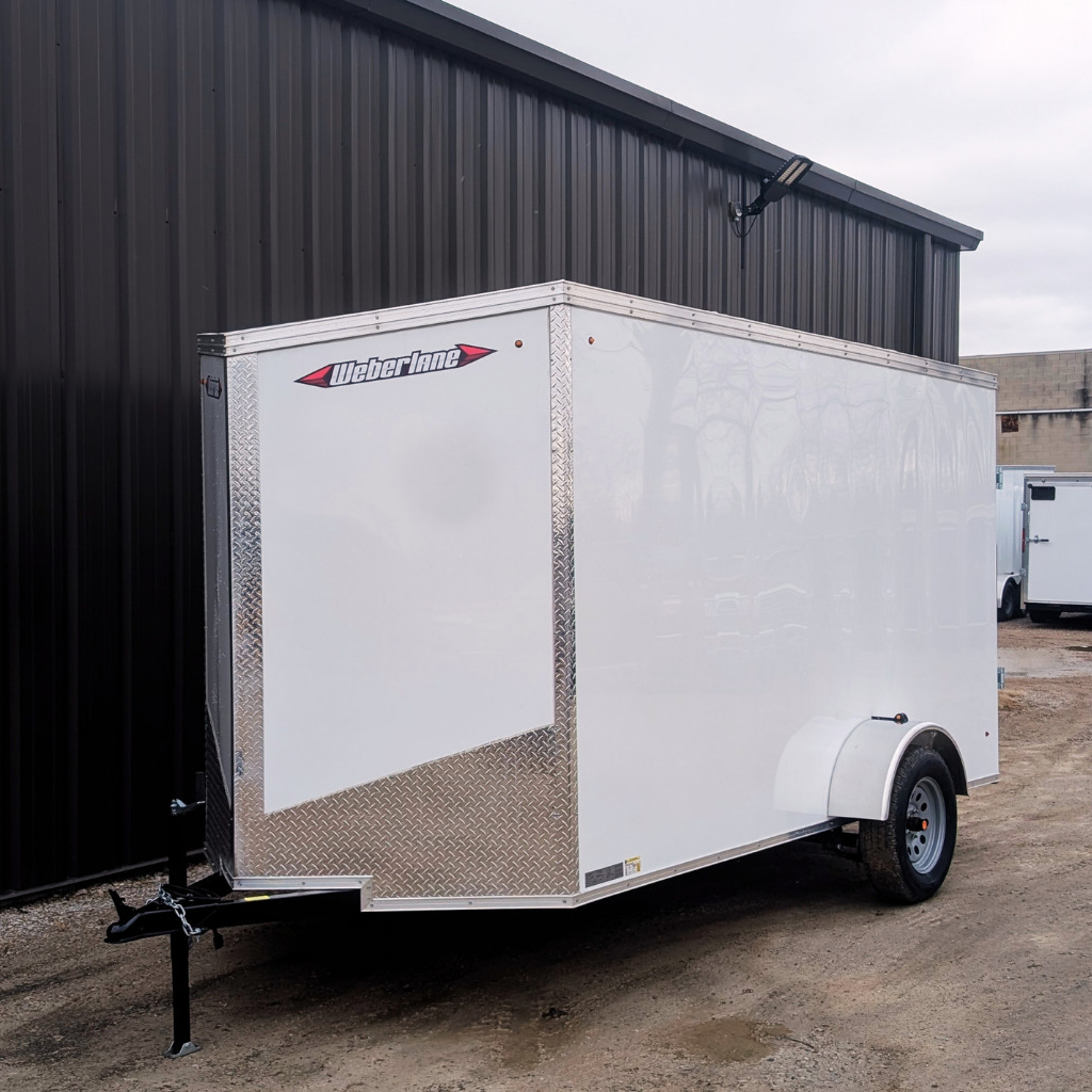 A 2026 WEBERLANE 6X12 Estate Series trailer with 6" extra height and ramp door in white is parked on gravel next to a dark metal building under cloudy skies.