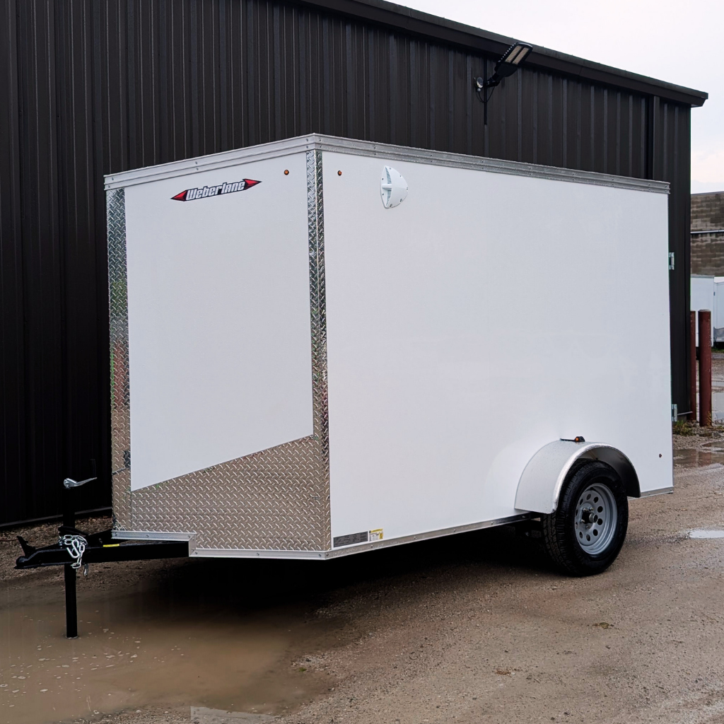A 2026 WEBERLANE 6X10 Estate Series white enclosed cargo trailer with 6" extra height and ramp door, single axle, is parked on wet gravel near a dark metal building.