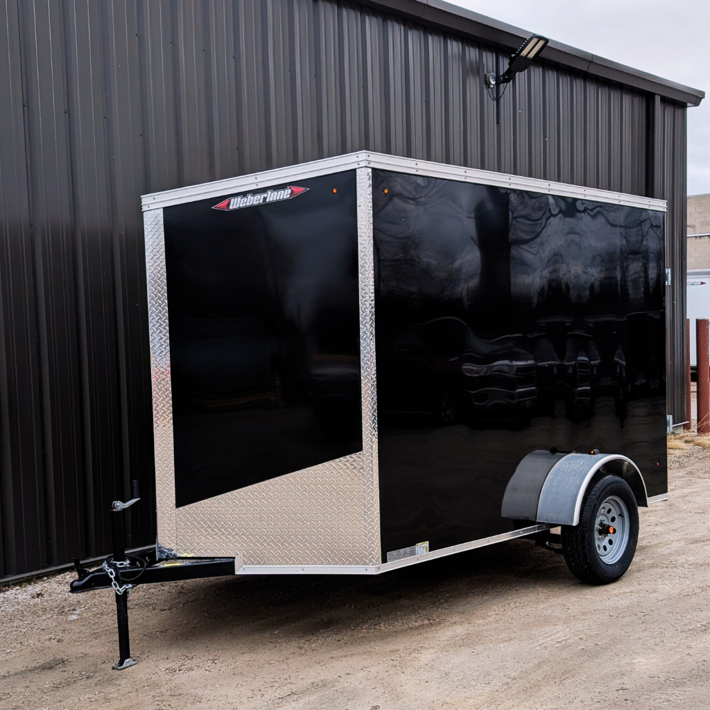 A 2026 WEBERLANE 6X10 Estate Series trailer with 6" extra height, ramp door, and black finish is parked on gravel beside a metal building.