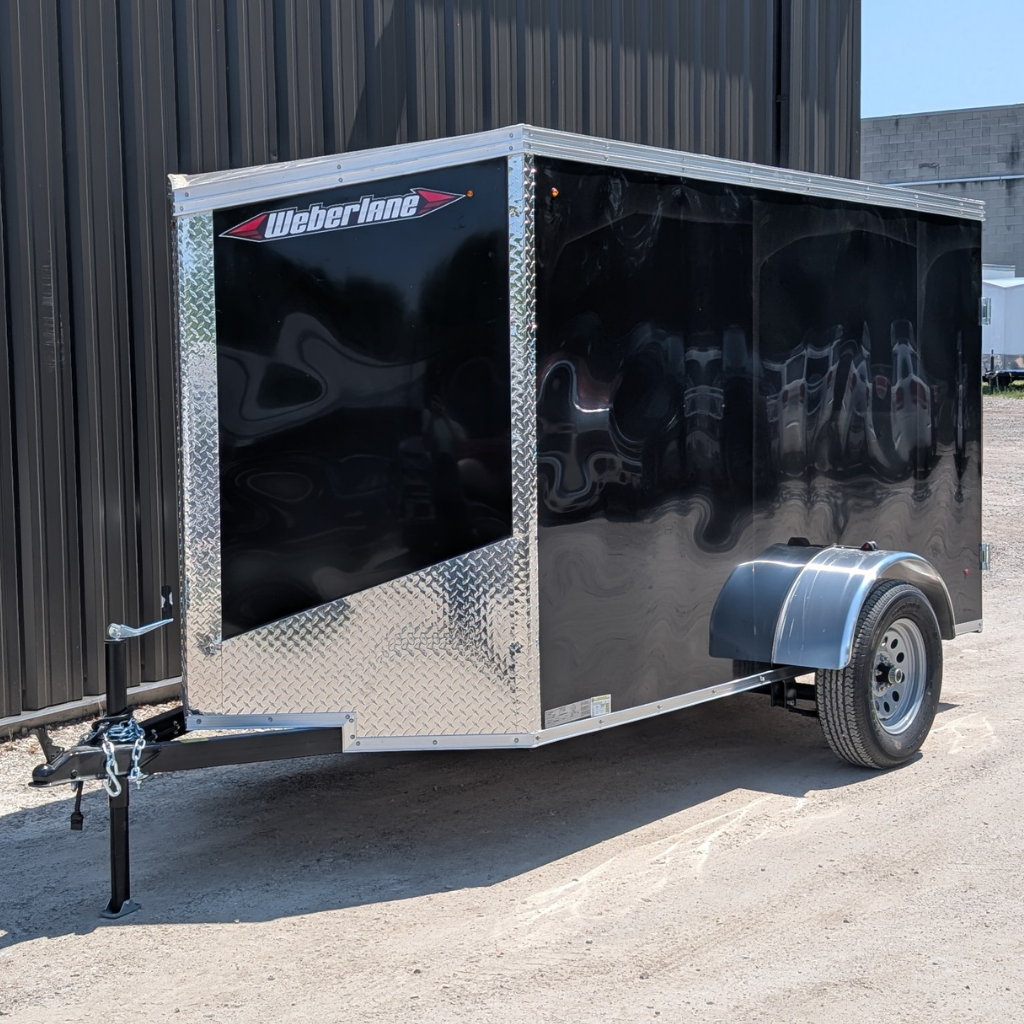 A 2026 WEBERLANE 5X10 Estate Series black enclosed utility trailer with 6" extra height and ramp door is parked on a gravel surface beside a dark-sided industrial building.
