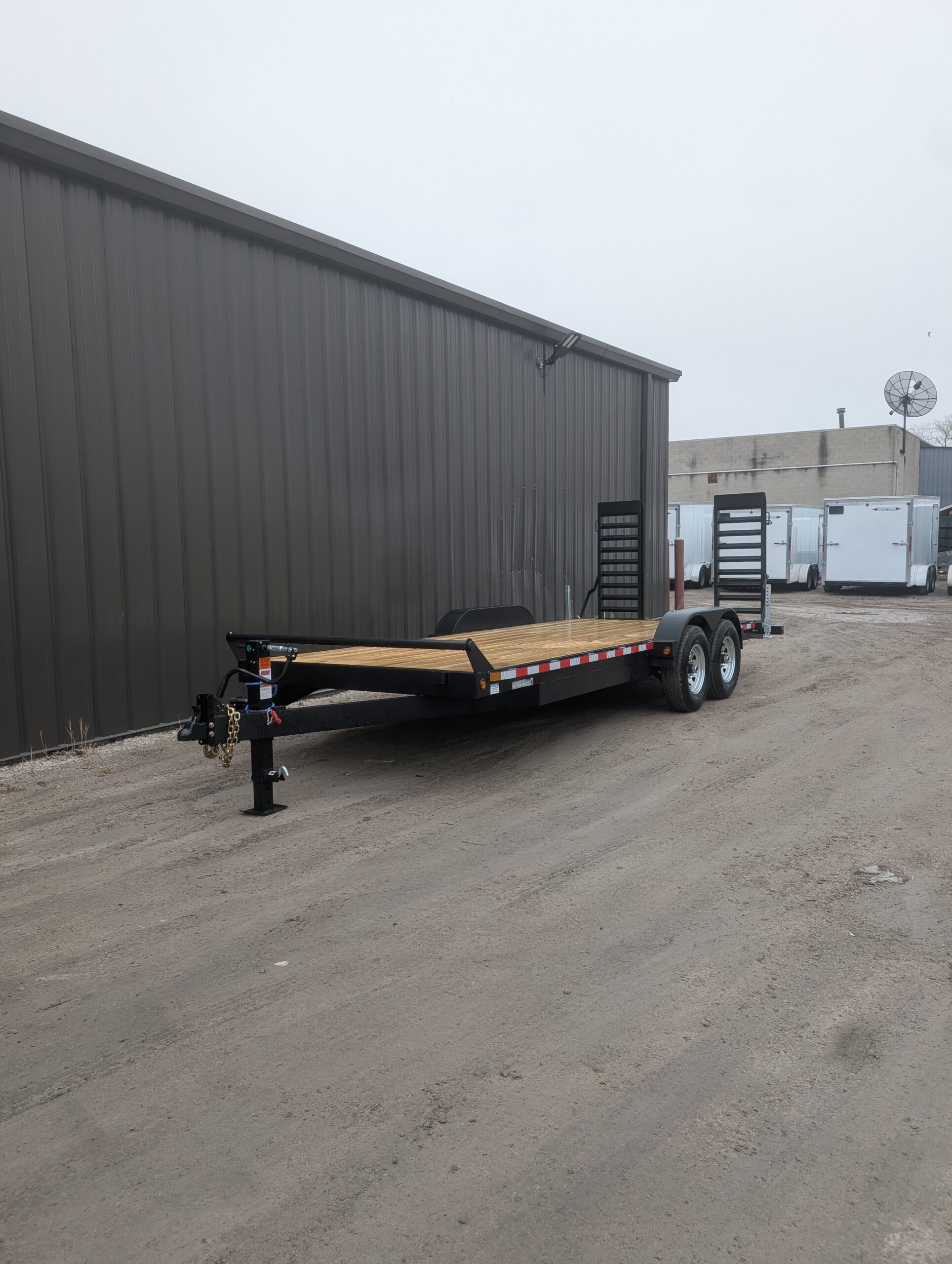 A 2026 CANADA TRAILER 7X20 CAR & EQUIPMENT 14000 GVWR flatbed utility trailer with a dual axle is parked beside a large metal building on a dirt lot.