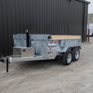 A 2026 WEBER LANE 6X10 Estate Series Dump Trailer (7000 GVWR, galvanized), featuring a wooden bed and metal toolbox, is parked on gravel beside a dark corrugated metal building.