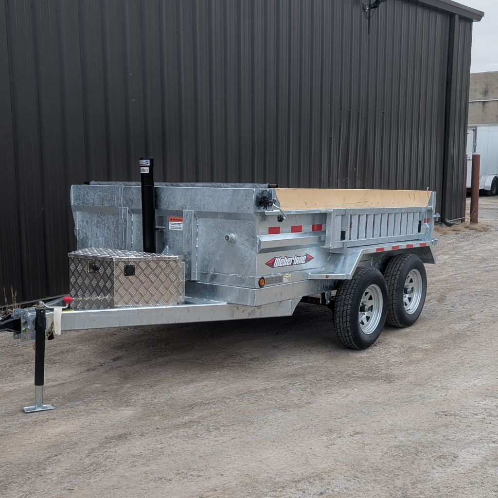 A 2026 WEBER LANE 6X10 Estate Series Dump Trailer (7000 GVWR, galvanized), featuring a wooden bed and metal toolbox, is parked on gravel beside a dark corrugated metal building.