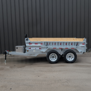 A WEBERLANE 6x10 ESTATE SERIES DUMP 10000 GVRW GALVANIZED ECDWL1072LP trailer with wood rail, dual axles, and reflective tape is parked on dirt in front of a brown metal building.