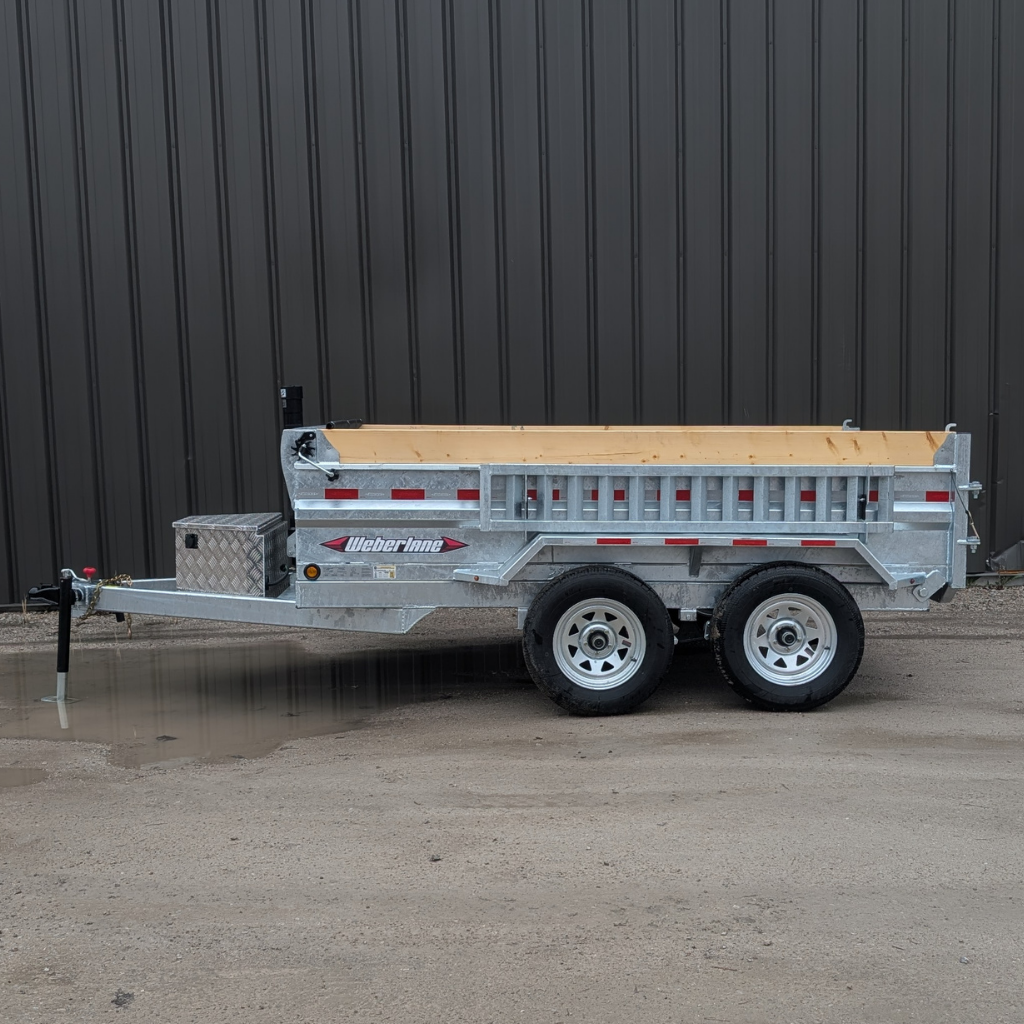 A WEBERLANE 6x10 ESTATE SERIES DUMP 10000 GVRW GALVANIZED ECDWL1072LP trailer with wood rail, dual axles, and reflective tape is parked on dirt in front of a brown metal building.
