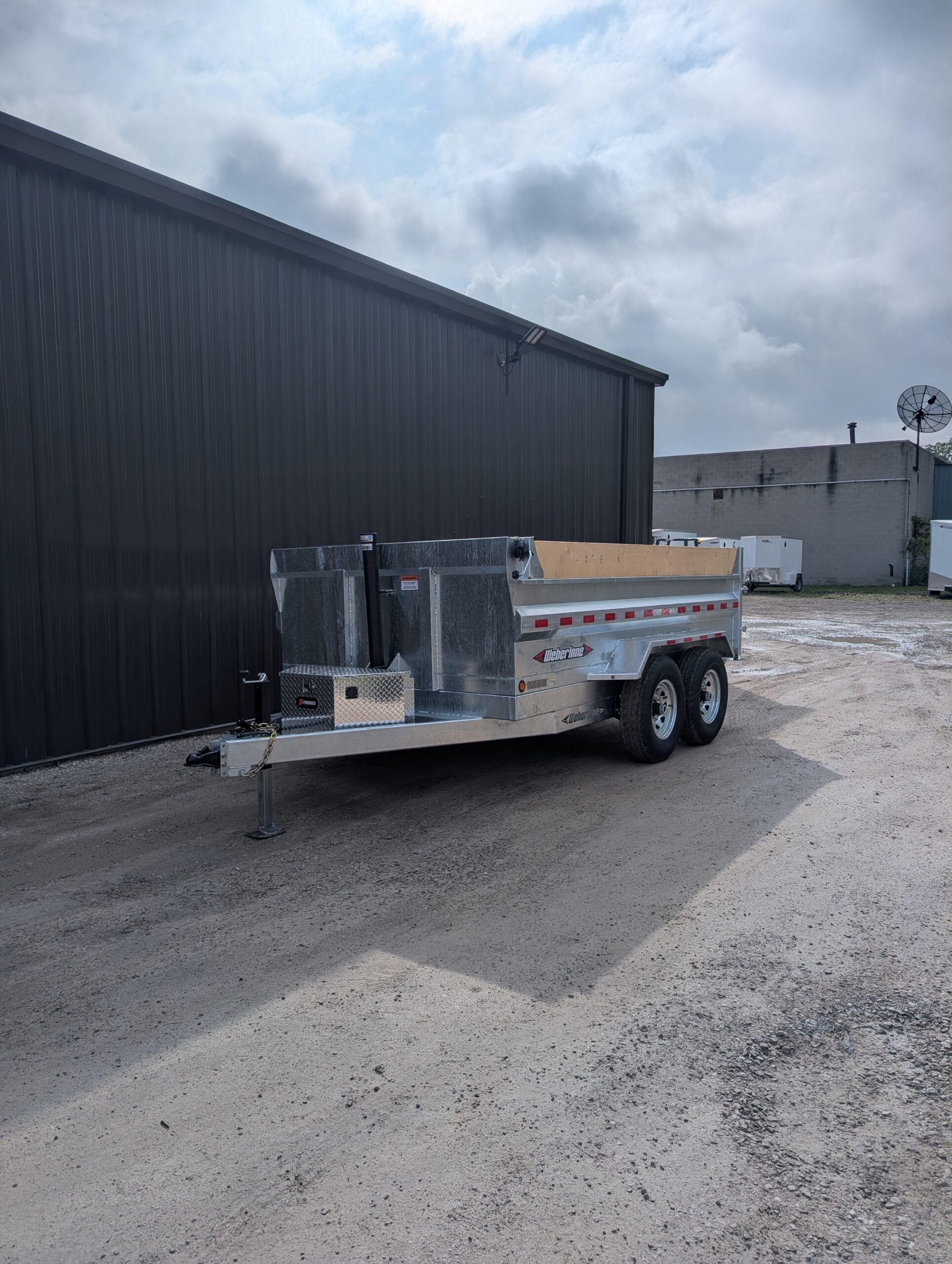 A USED DWL1272LP 2025 WEBER LANE 82X12 CONTRACTOR SERIES DUMP 14,000 GVWR GALVANIZED trailer is parked on gravel beside a large black metal building.
